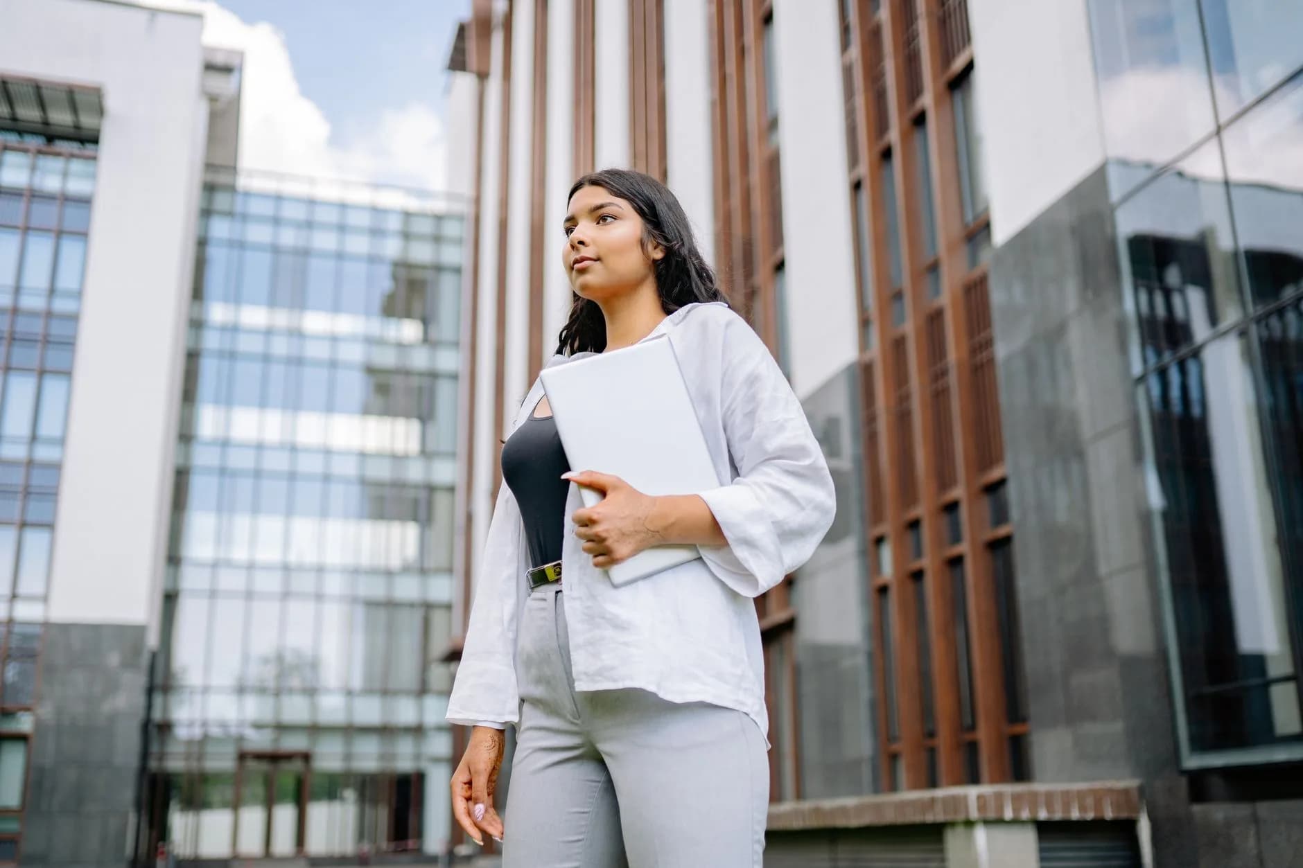 Confident businesswoman stands holding a laptop in front of a modern building.