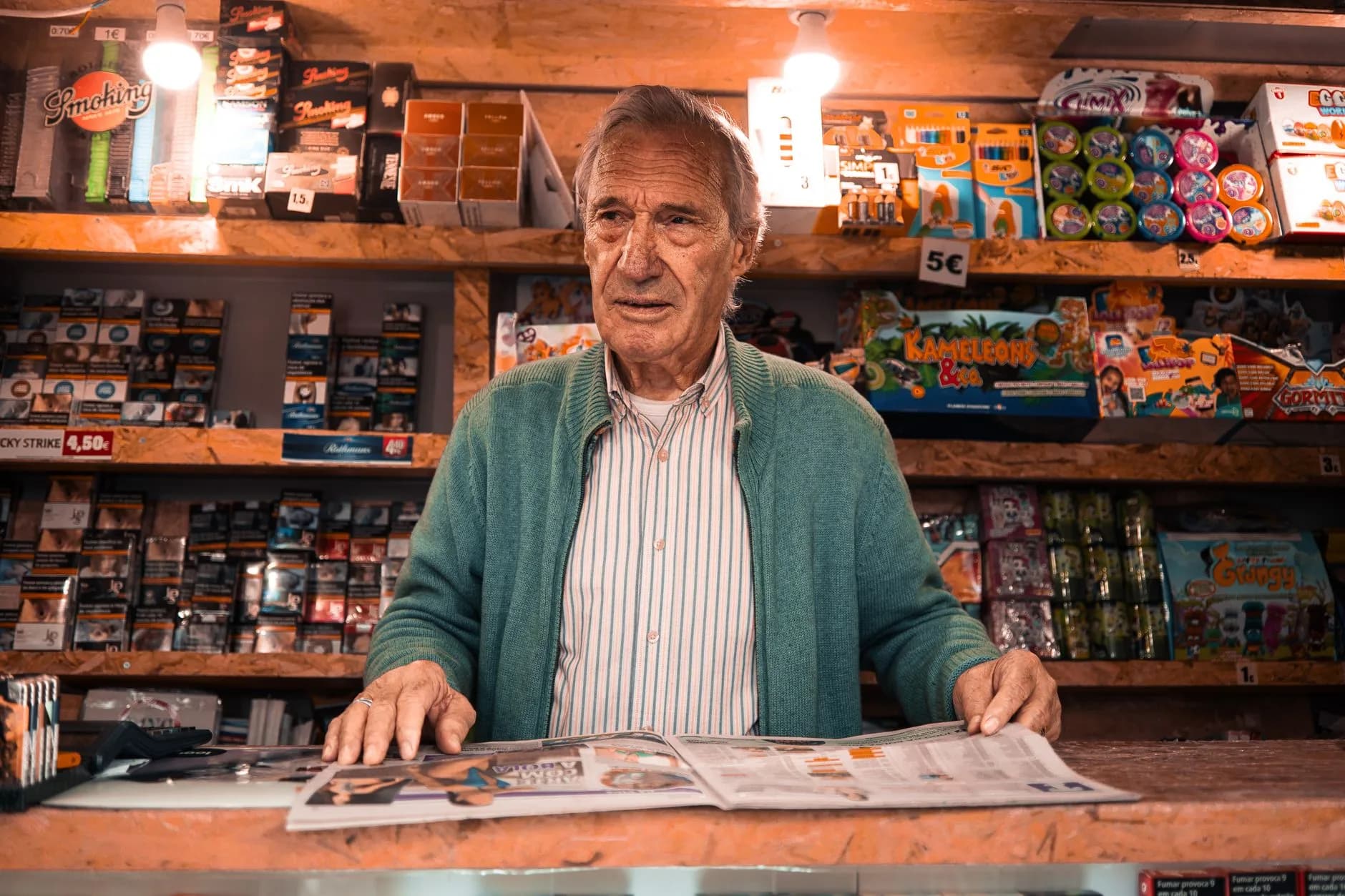 An elderly shopkeeper stands behind the counter in a Lisbon newsstand, surrounded by colorful products.