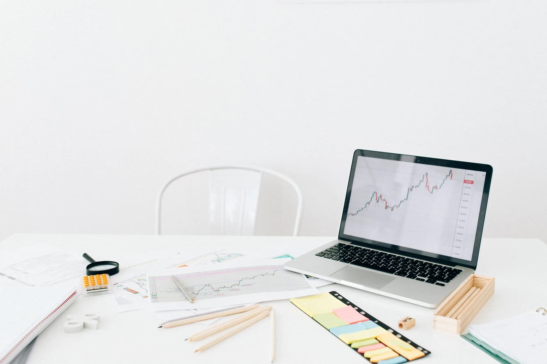 A modern workspace featuring a laptop displaying financial stock charts with documents and stationery on a white desk.