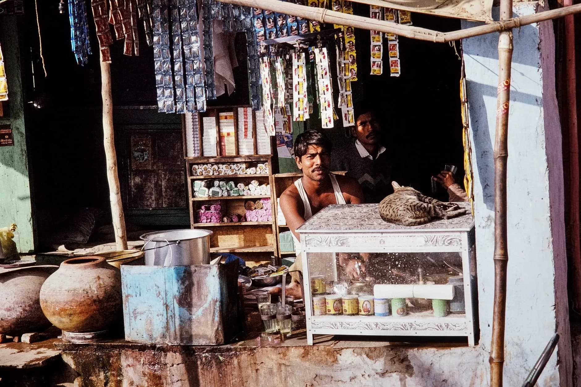 Rustic Indian shop with people and a cat in a village setting, showcasing traditional merchandise.
