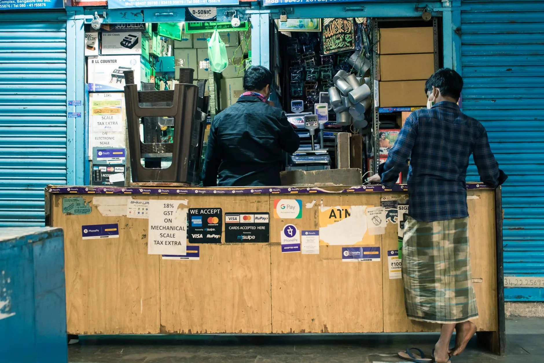 Two men at a shop counter in Bengaluru marketplace with payment signs and products displayed.