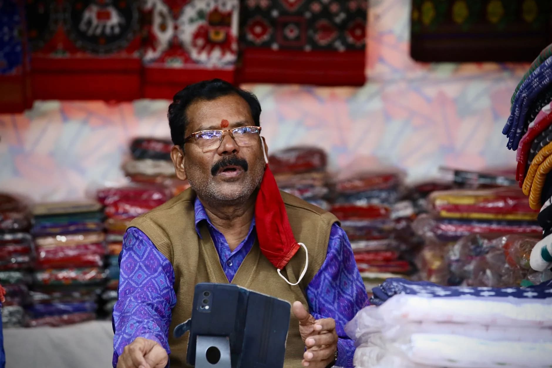 Man selling traditional fabrics at a colorful market stall, engaging with a customer.
