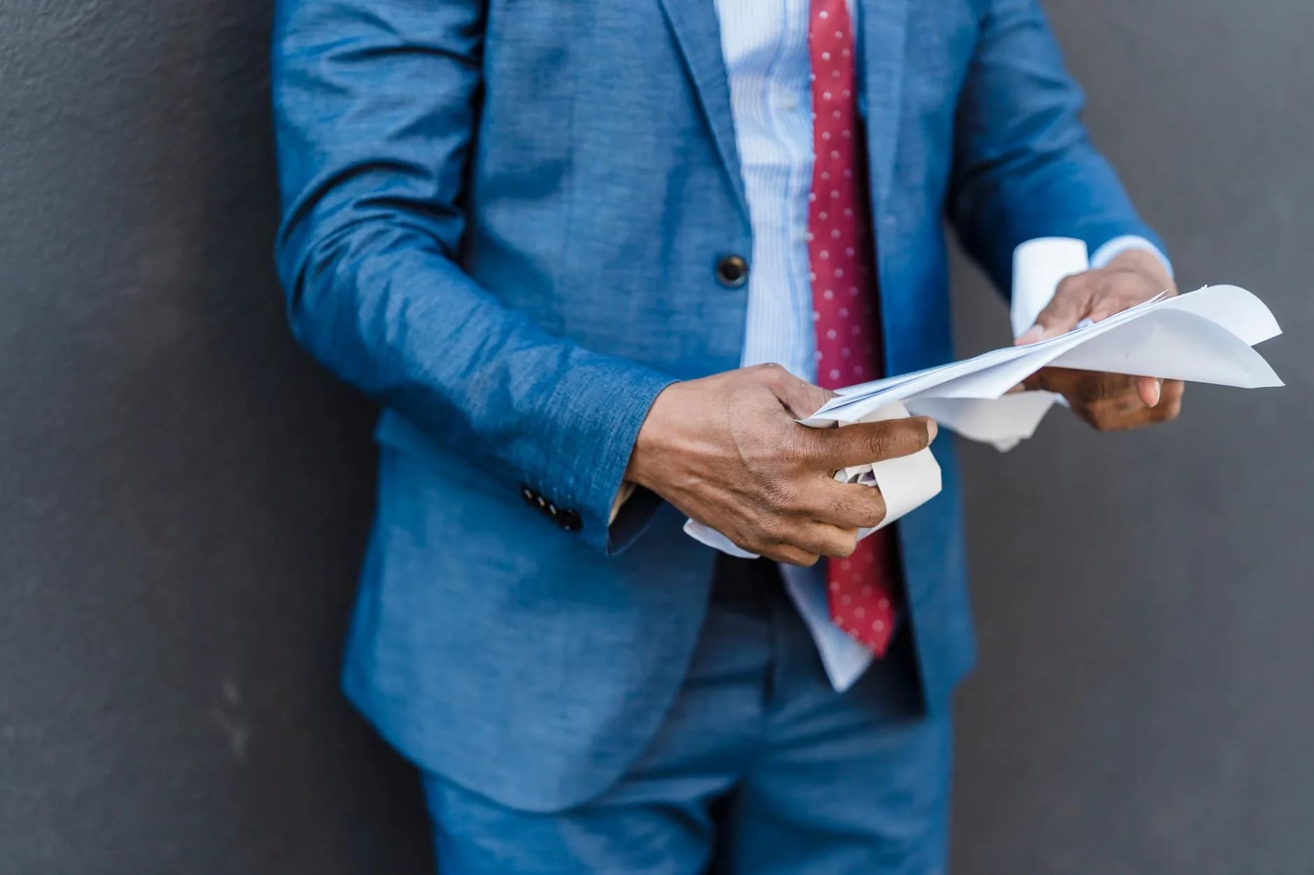 Crop unrecognizable male entrepreneur in formal suit reading documents in hands while standing on gray background in cit